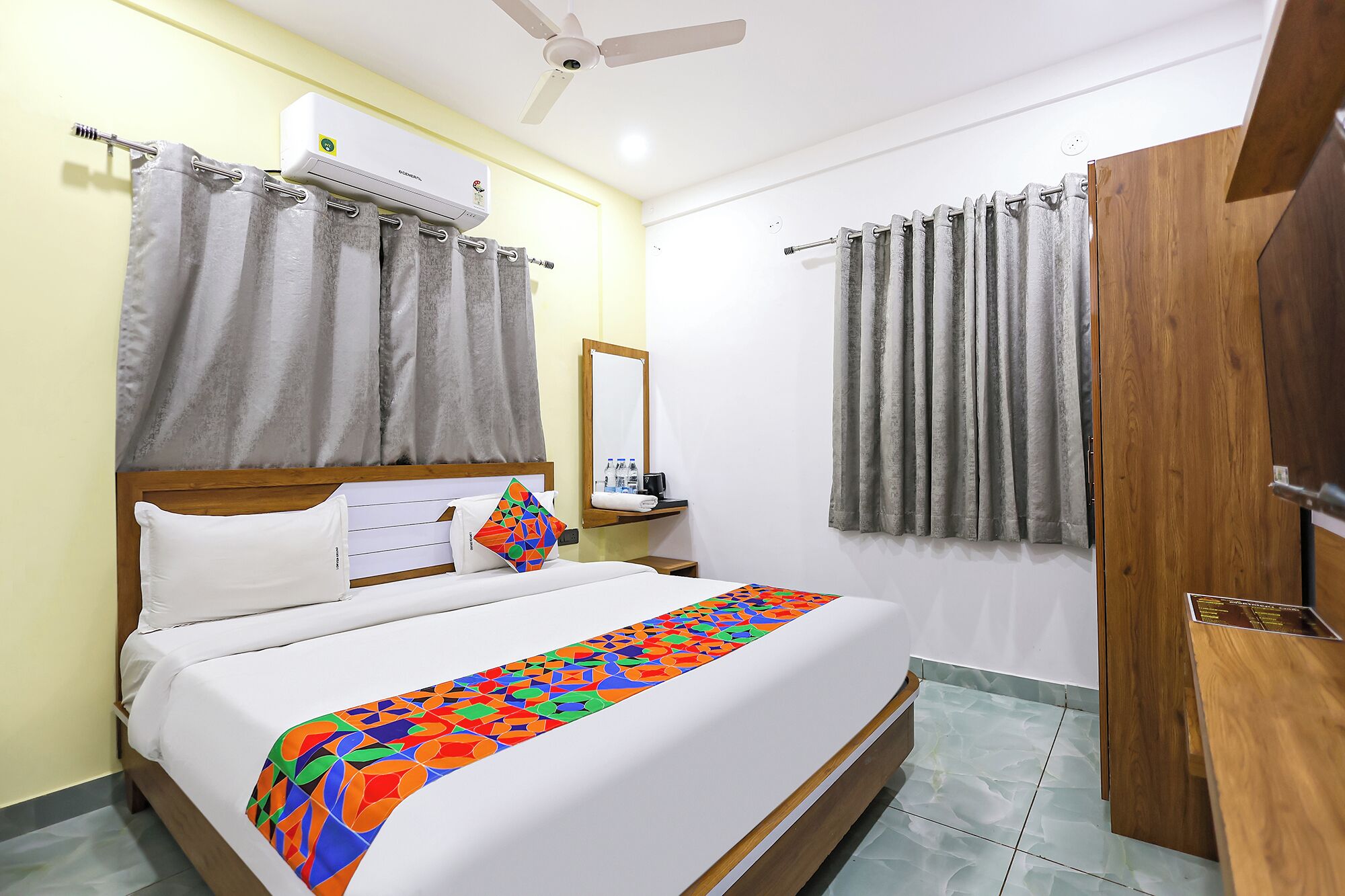 A hotel room with a bed, curtains, a ceiling fan, a wooden wardrobe, and a bathroom mirror.