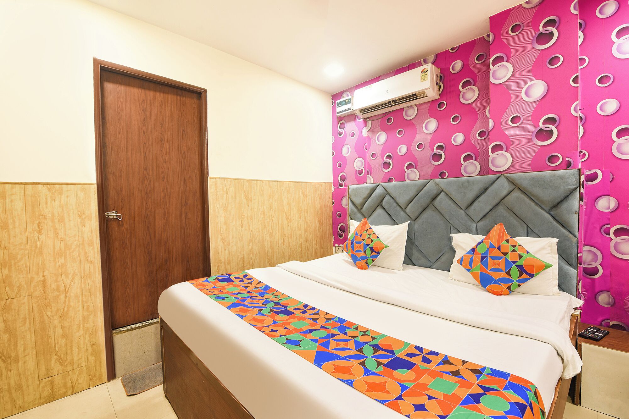 A bedroom with a wooden door, a bed with a patterned bedspread, pillows, a headboard, and a wall air conditioner.