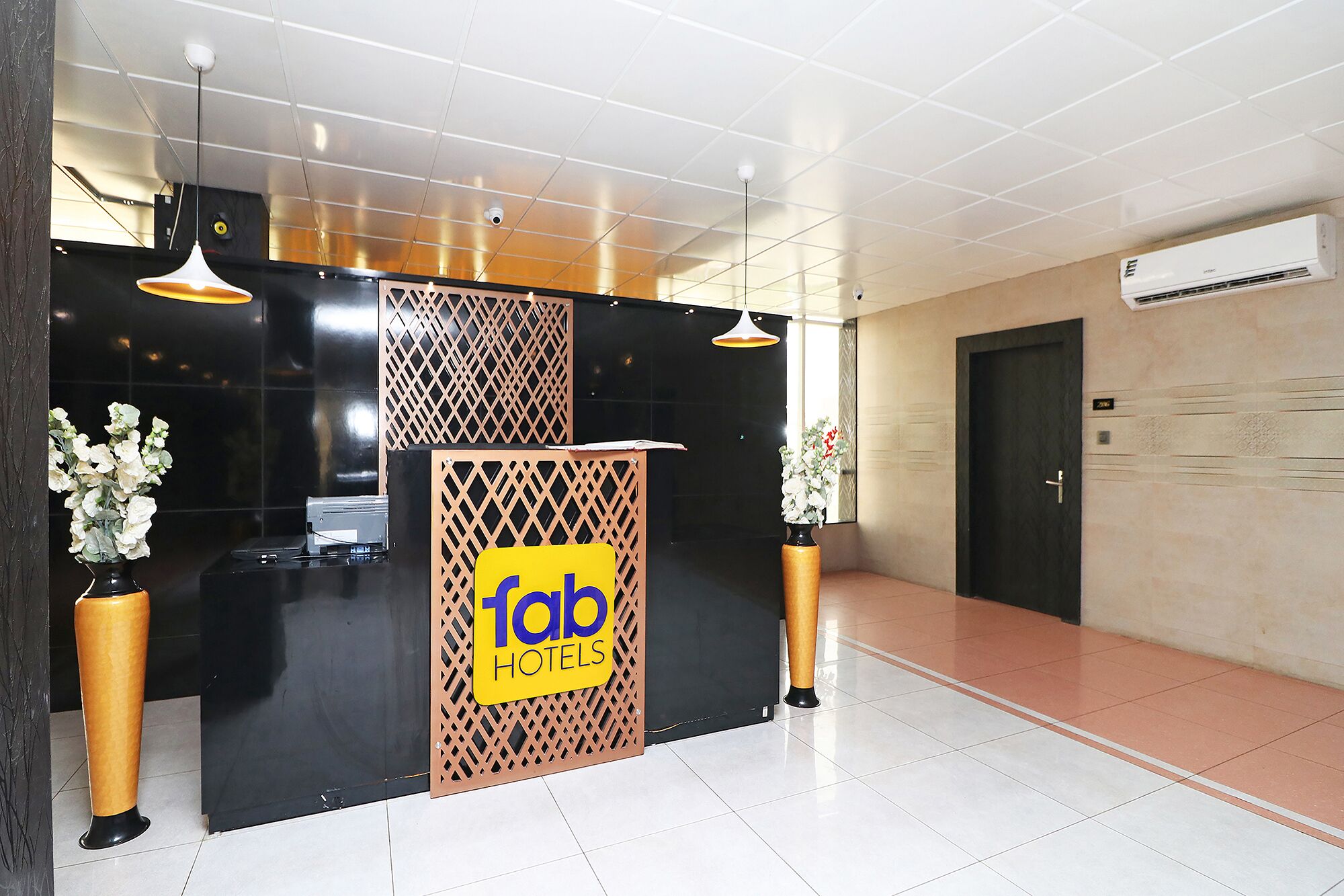 Un comptoir d’accueil avec une enseigne indiquant « fab hotels », un comptoir noir, deux plantes en pot et un sol carrelé.