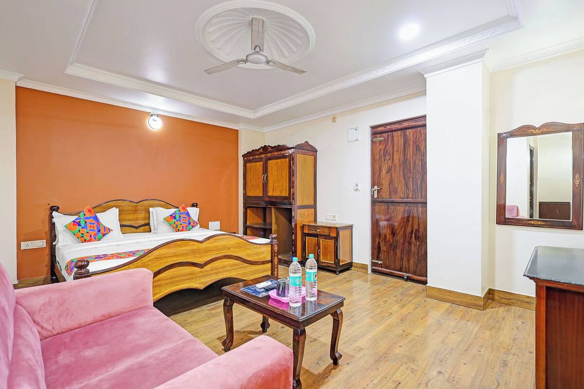 A bedroom with a bed, a wooden wardrobe, a small table with bottles, a pink chair, and a ceiling fan.