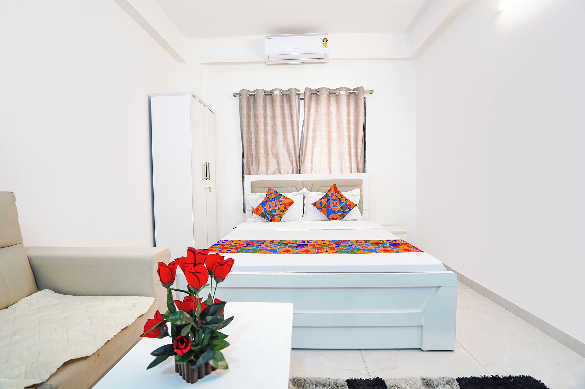A bedroom with a bed, a bedside table with a vase of flowers, a sofa, a window with curtains, and an air conditioning unit.