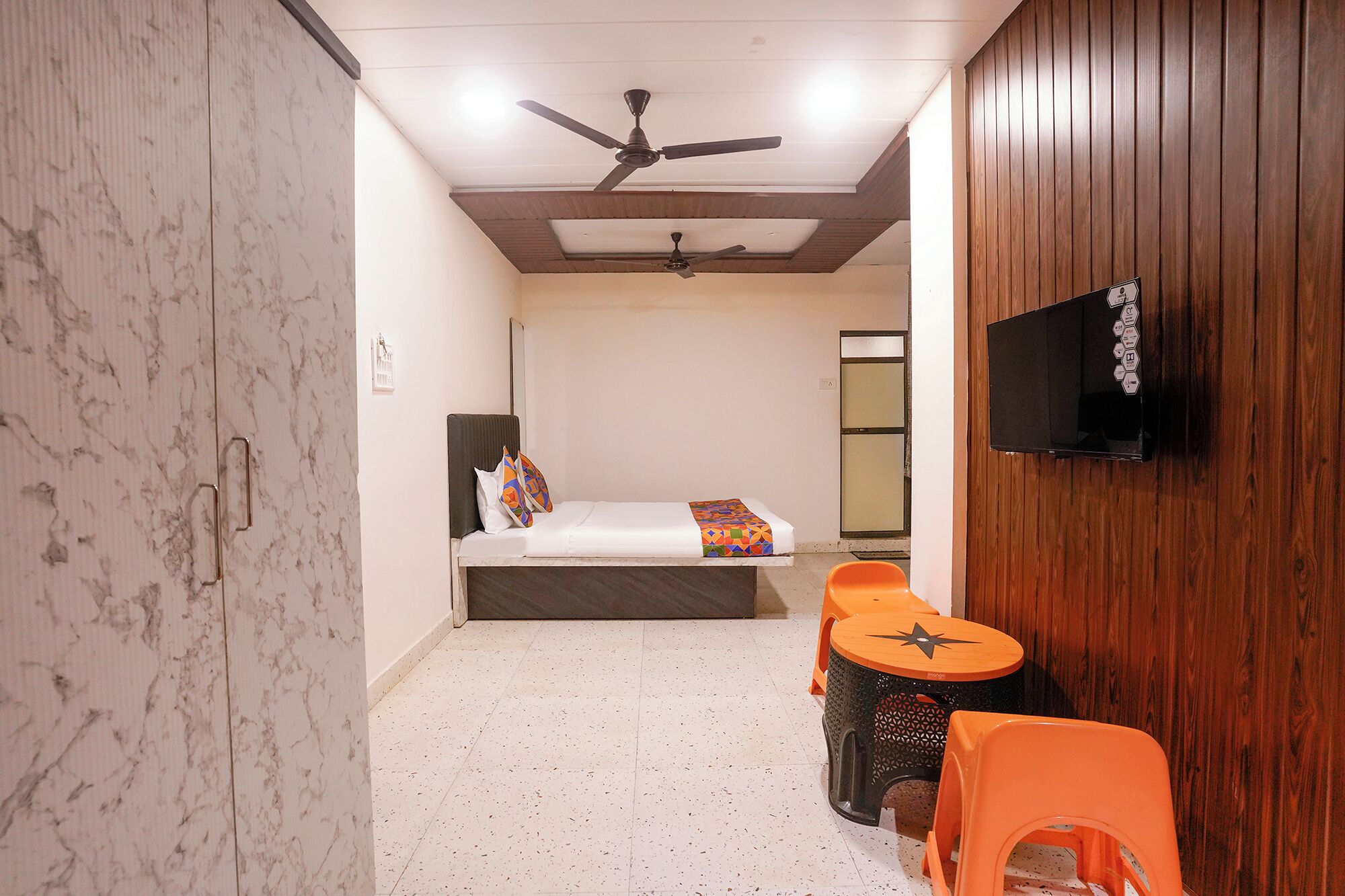 Een hotelkamer met een bed, een plafondventilator, een aan de muur bevestigde televisie en twee oranje stoelen.