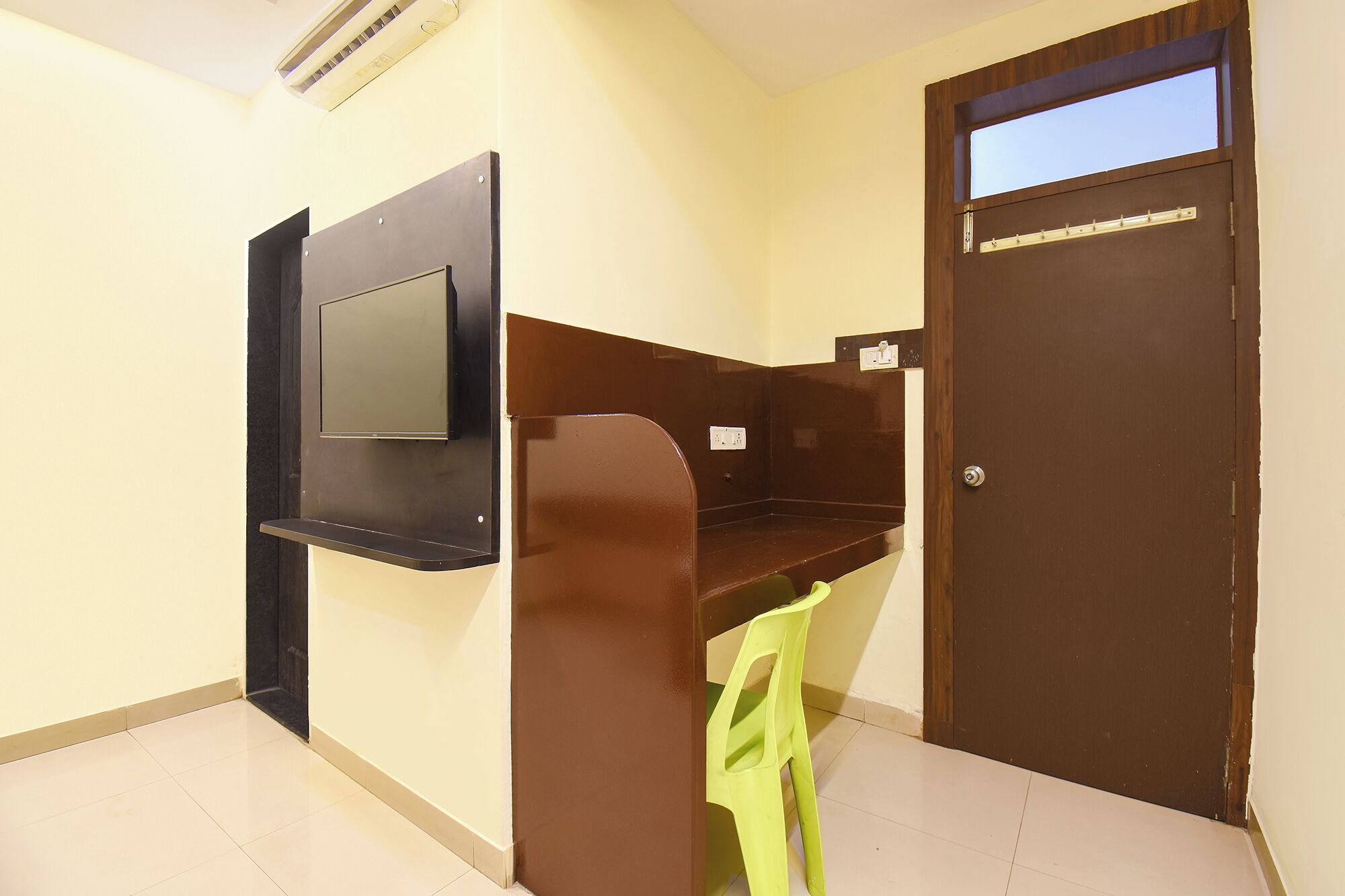 Una habitación con una televisión montada en la pared, una puerta con ventanal y una silla de plástico verde.