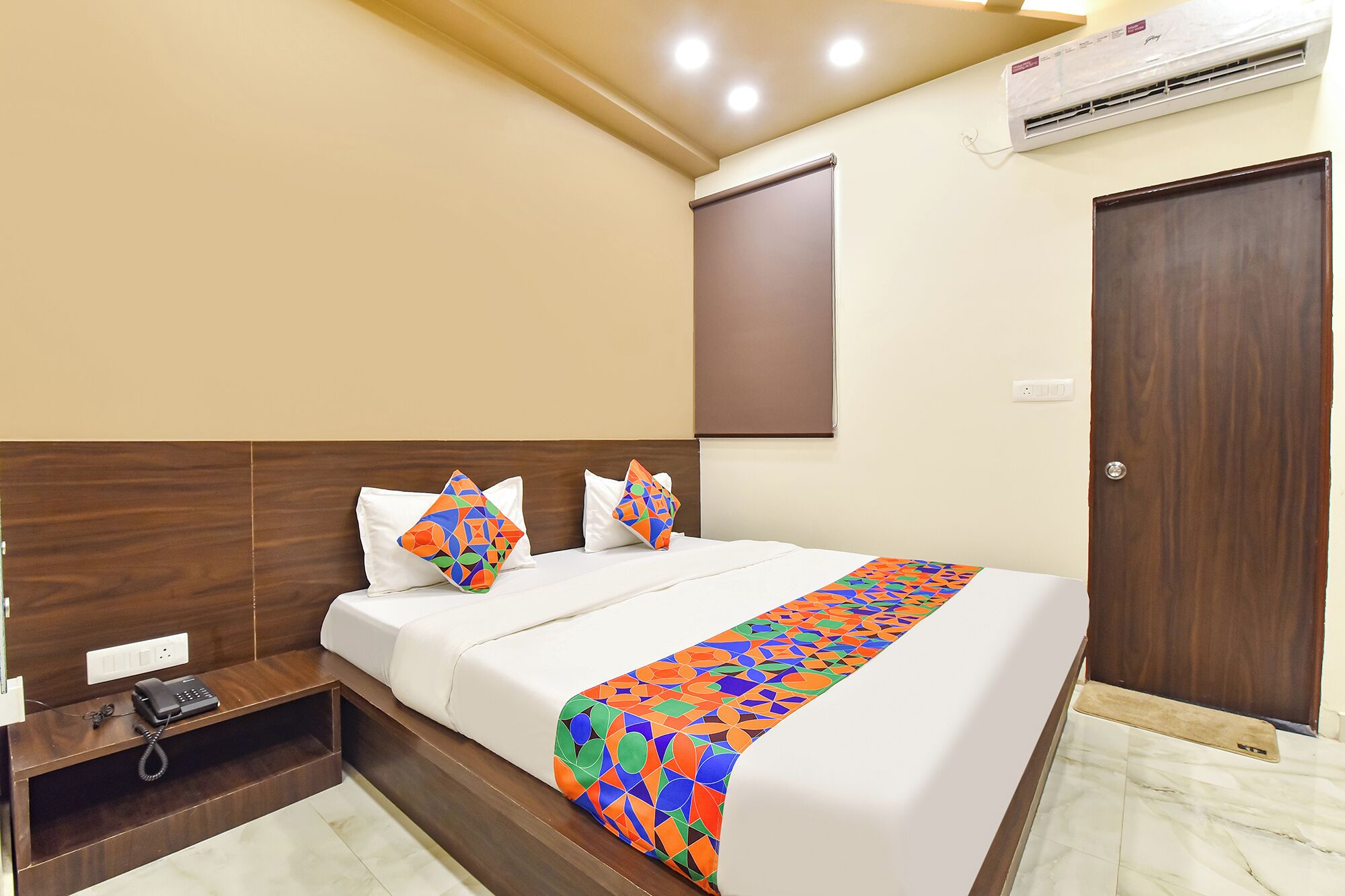 A hotel room with a bed, a bedside phone, a wall-mounted air conditioner, and a door.