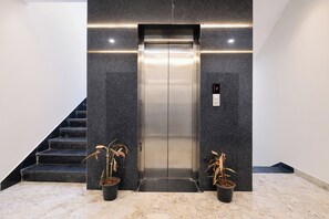 An elevator with a digital display, flanked by two potted plants, in a modern lobby with marble flooring and a staircase.