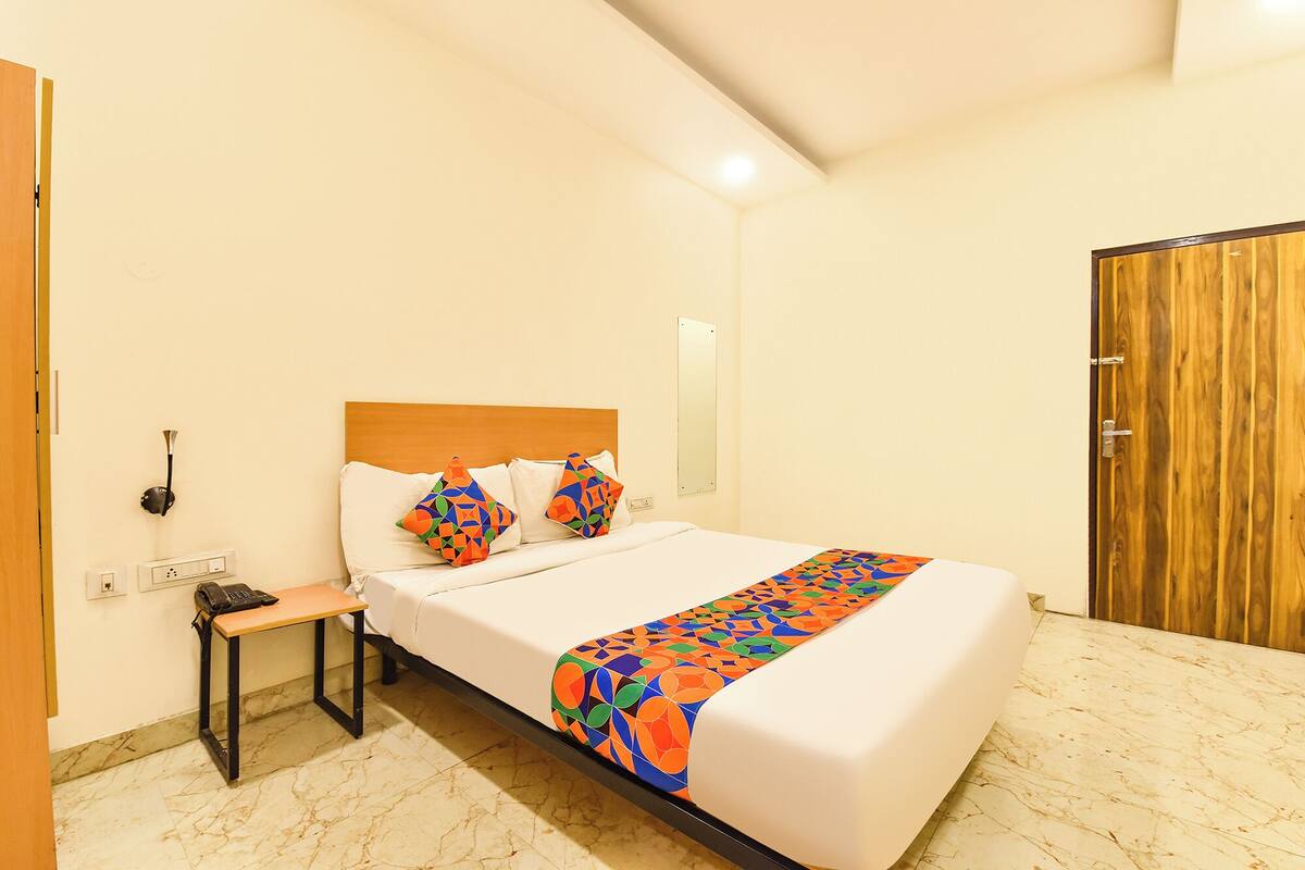 A hotel room with a bed, a bedside table with a phone, a wall-mounted lamp, and a wooden door.