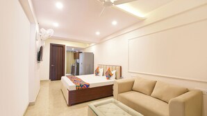 Habitación de hotel con cama, mesita de vidrio, sofá color beige, ventilador de techo y minibar.