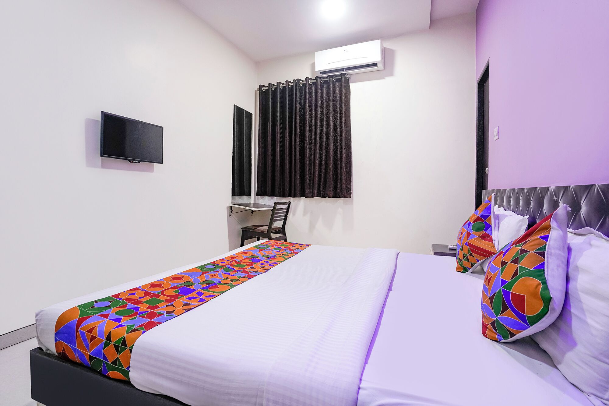 A hotel room with a bed, a television, an air conditioning unit, a chair, and a window with curtains.