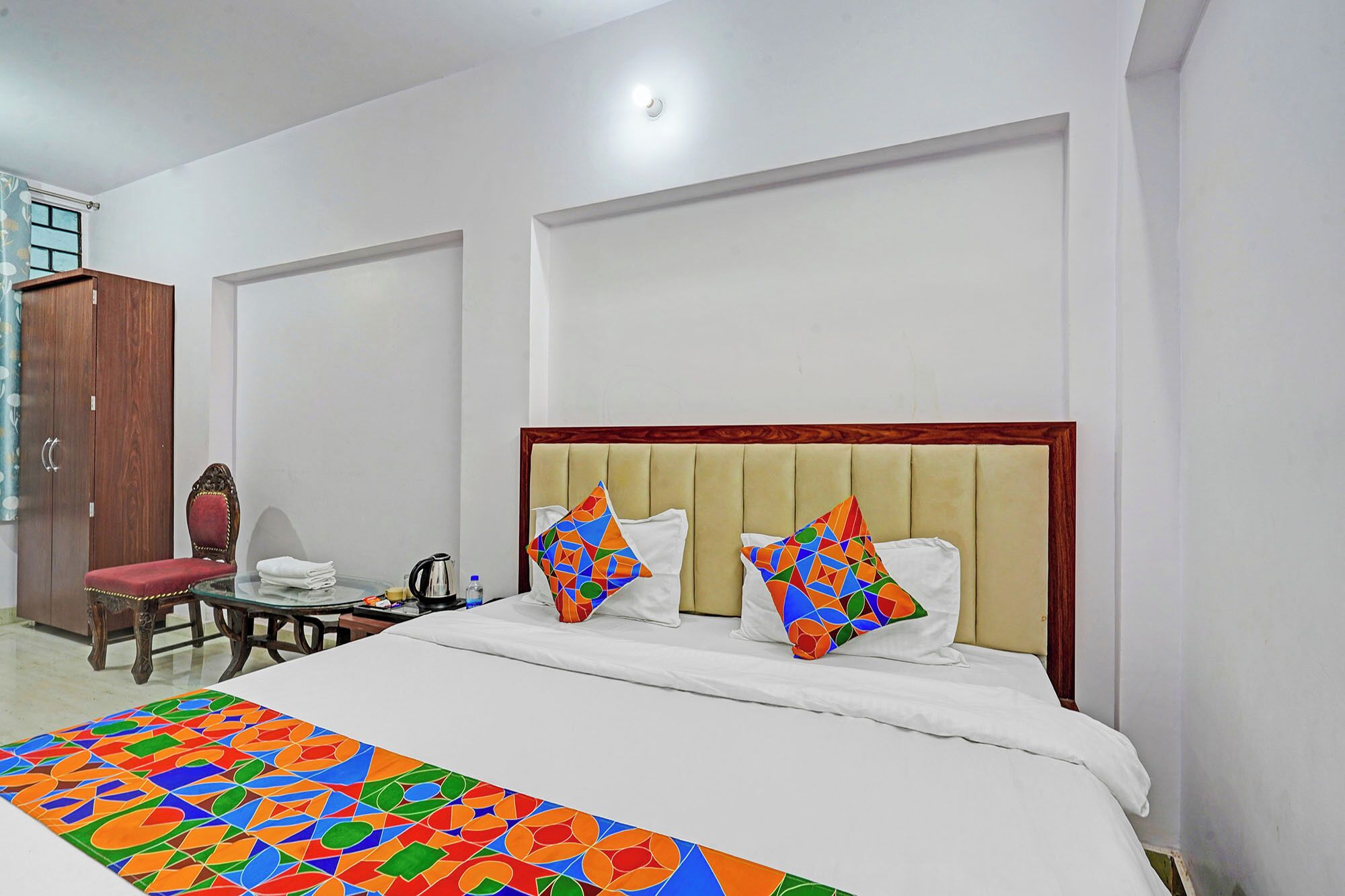 A hotel room with a bed, a wooden wardrobe, a chair, and a table with a kettle.