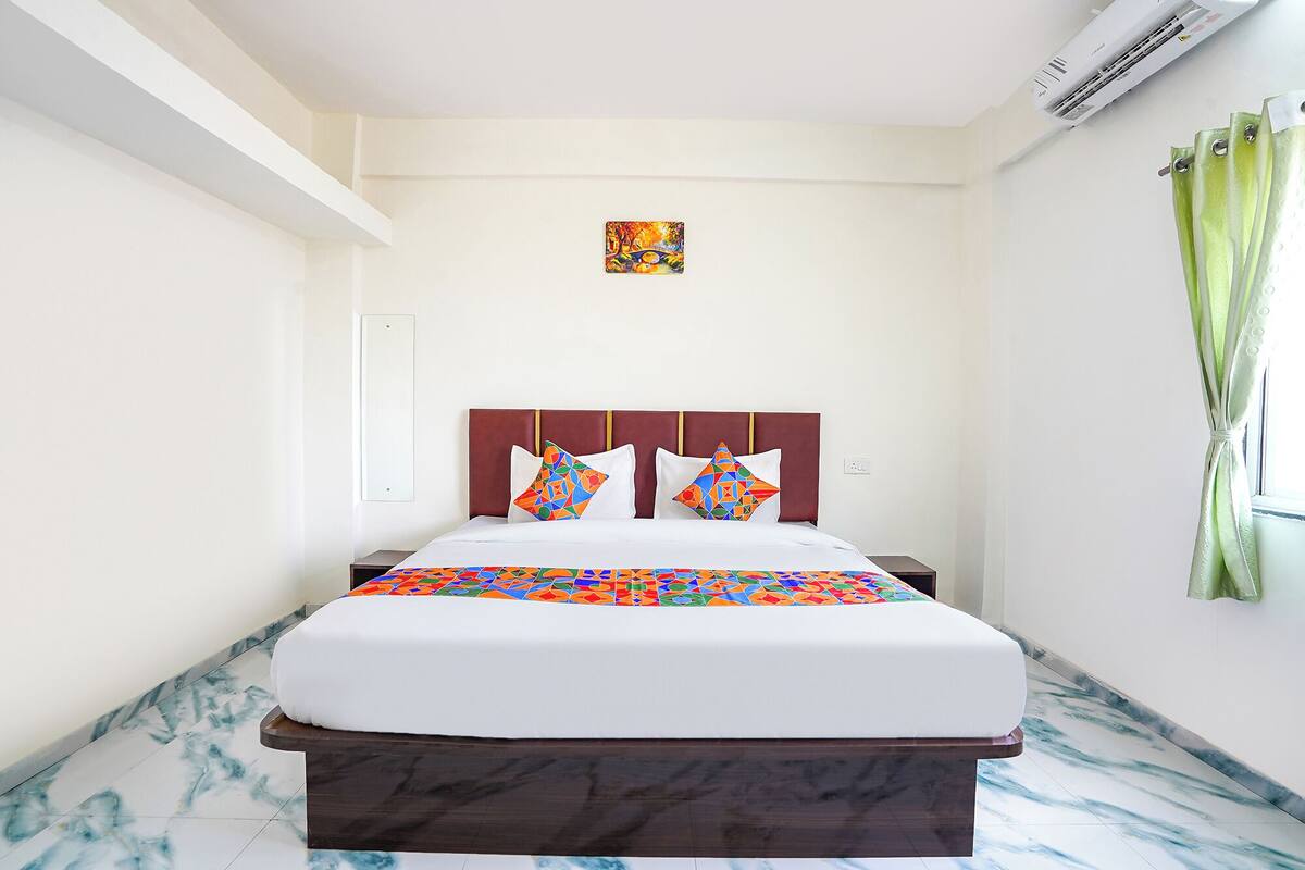 A hotel room with a bed, a headboard, a painting, a window with curtains, and an air conditioning unit.