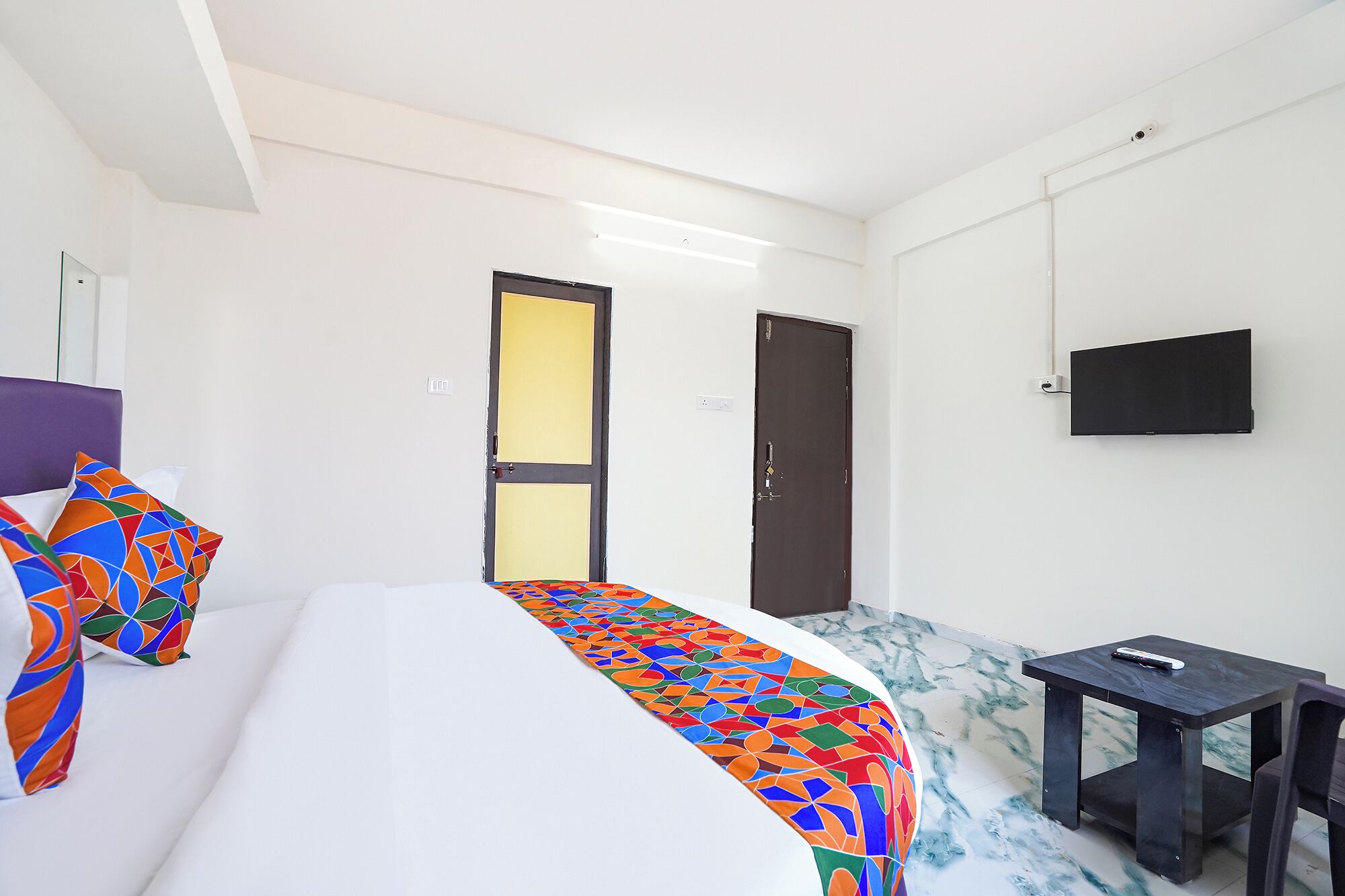 Una habitación de hotel con cama, una mesita pequeña, televisor montado en la pared y dos puertas.