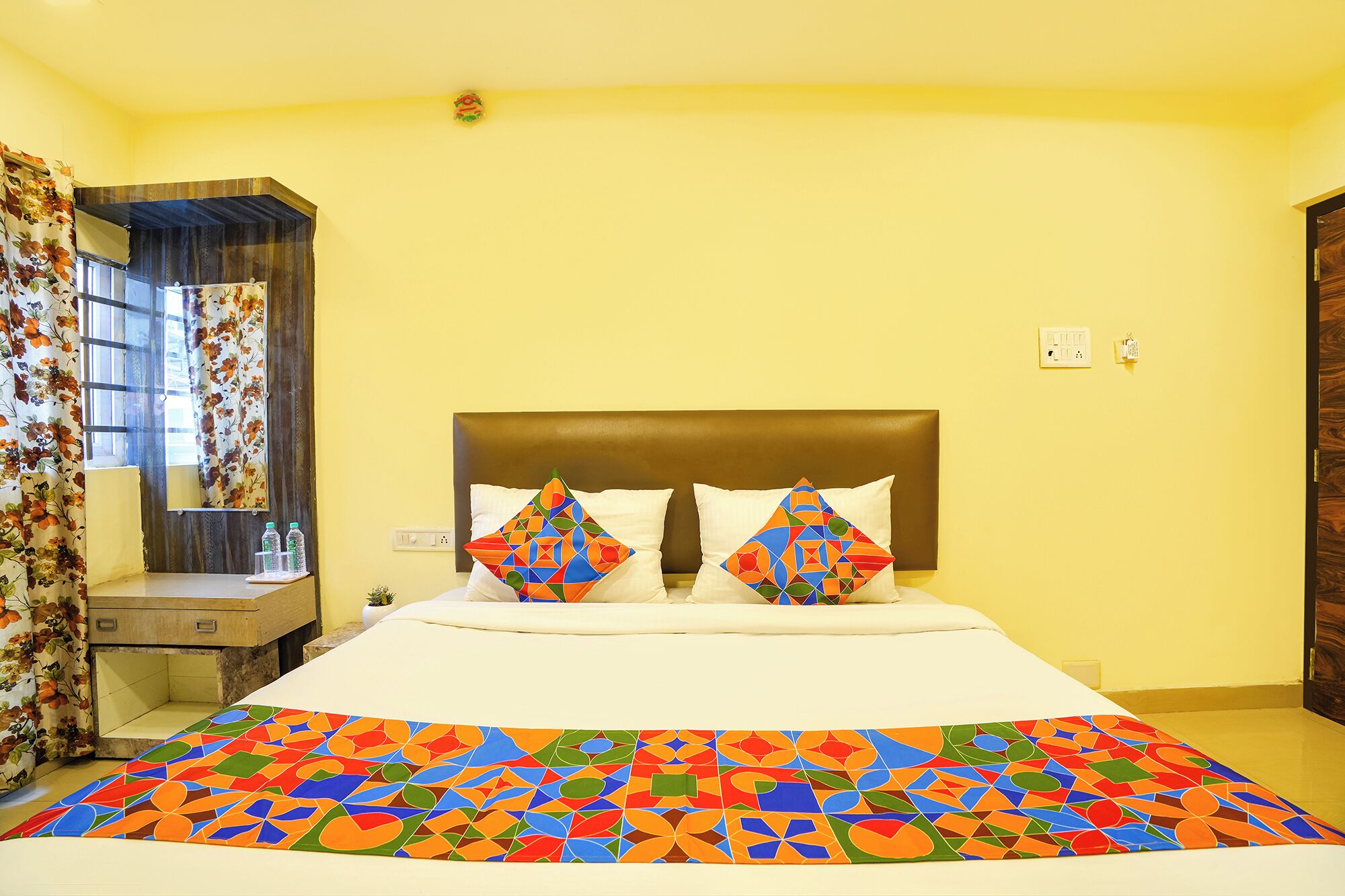 A hotel room with a bed, a nightstand, a window with curtains, and a wall-mounted light fixture.