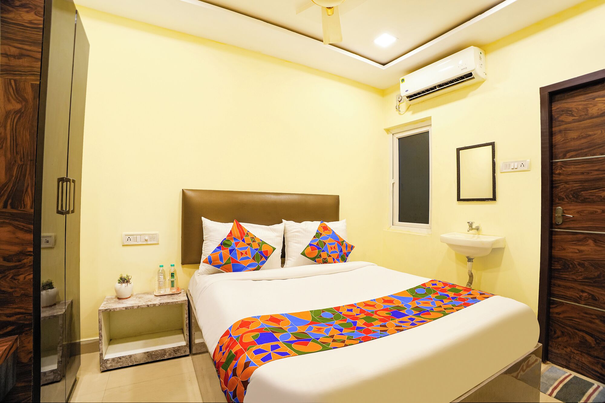 A hotel room with a bed, bedside table, nightstand, wall-mounted air conditioning unit, window, and a mirror.