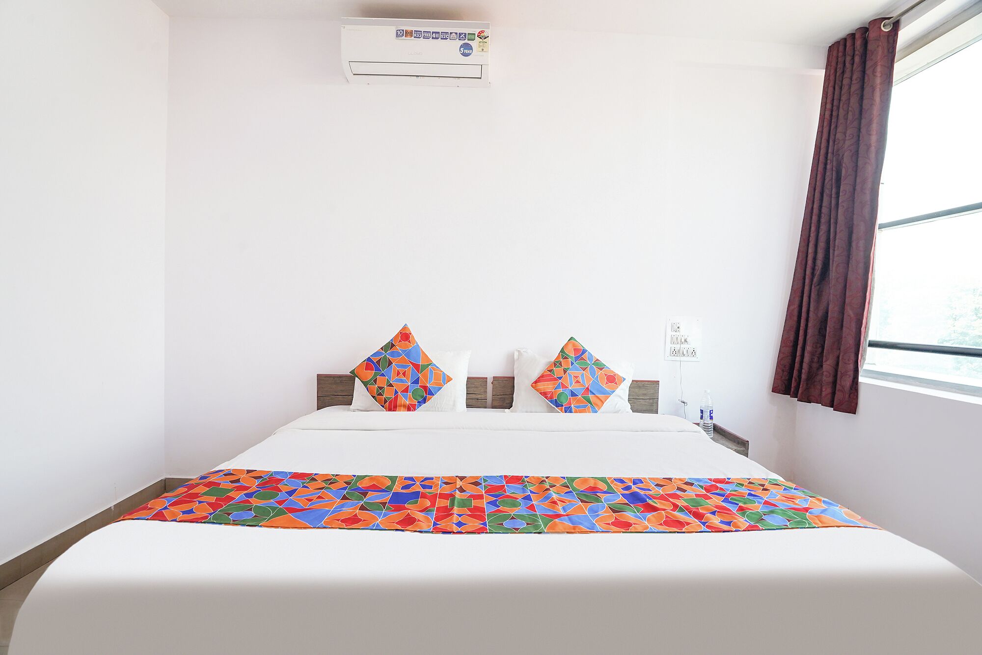 A hotel room with a bed, pillows, a window with curtains, and an air conditioning unit.