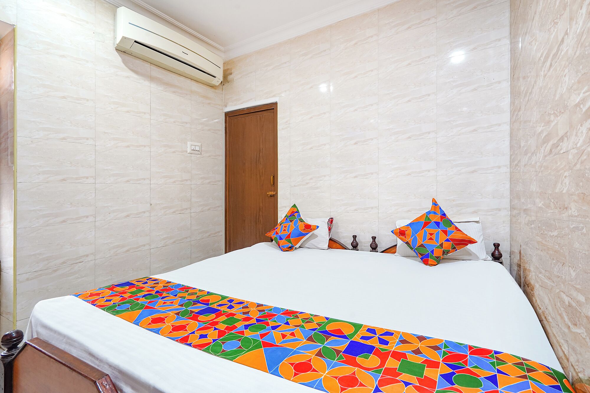 Una habitación de hotel con una cama, una puerta de madera, un equipo de aire acondicionado y una pared con azulejos.