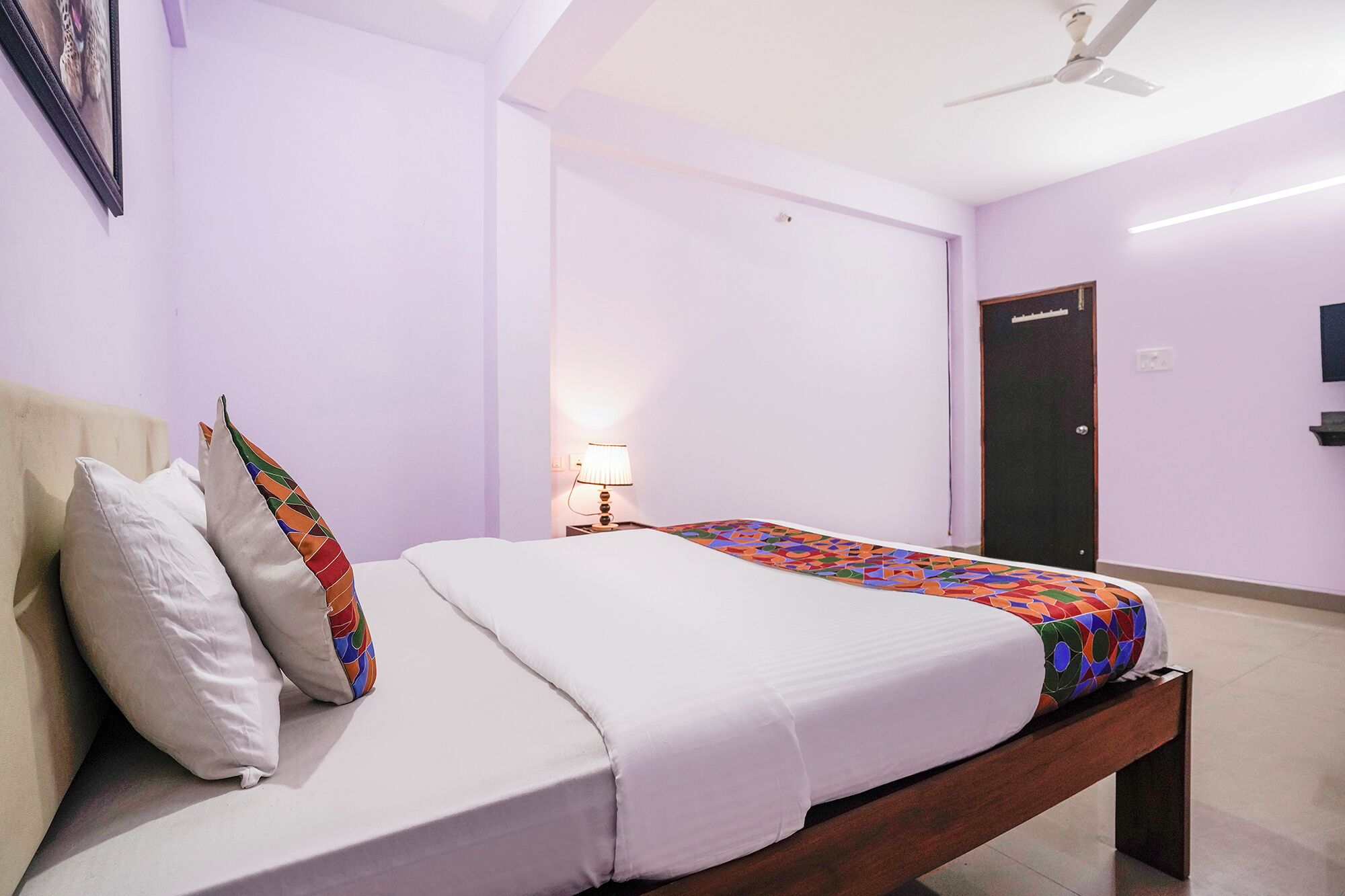 A single bed with a white mattress and colorful blanket, a bedside table with a lamp, a ceiling fan, and a door in a room with light purple walls.