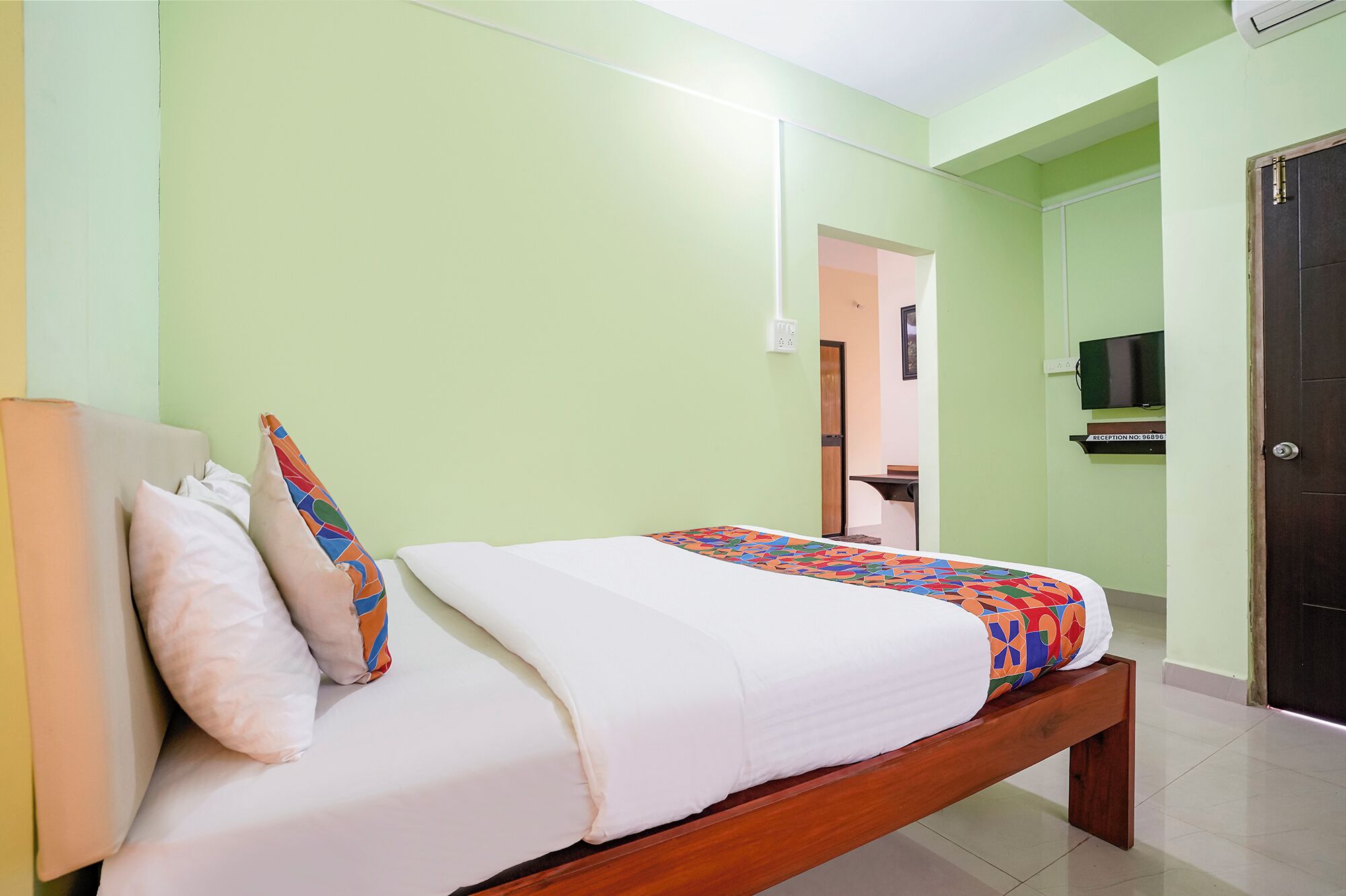 A single bed with a wooden frame, white bedding, and colorful pillows. A television is mounted on the wall to the right.