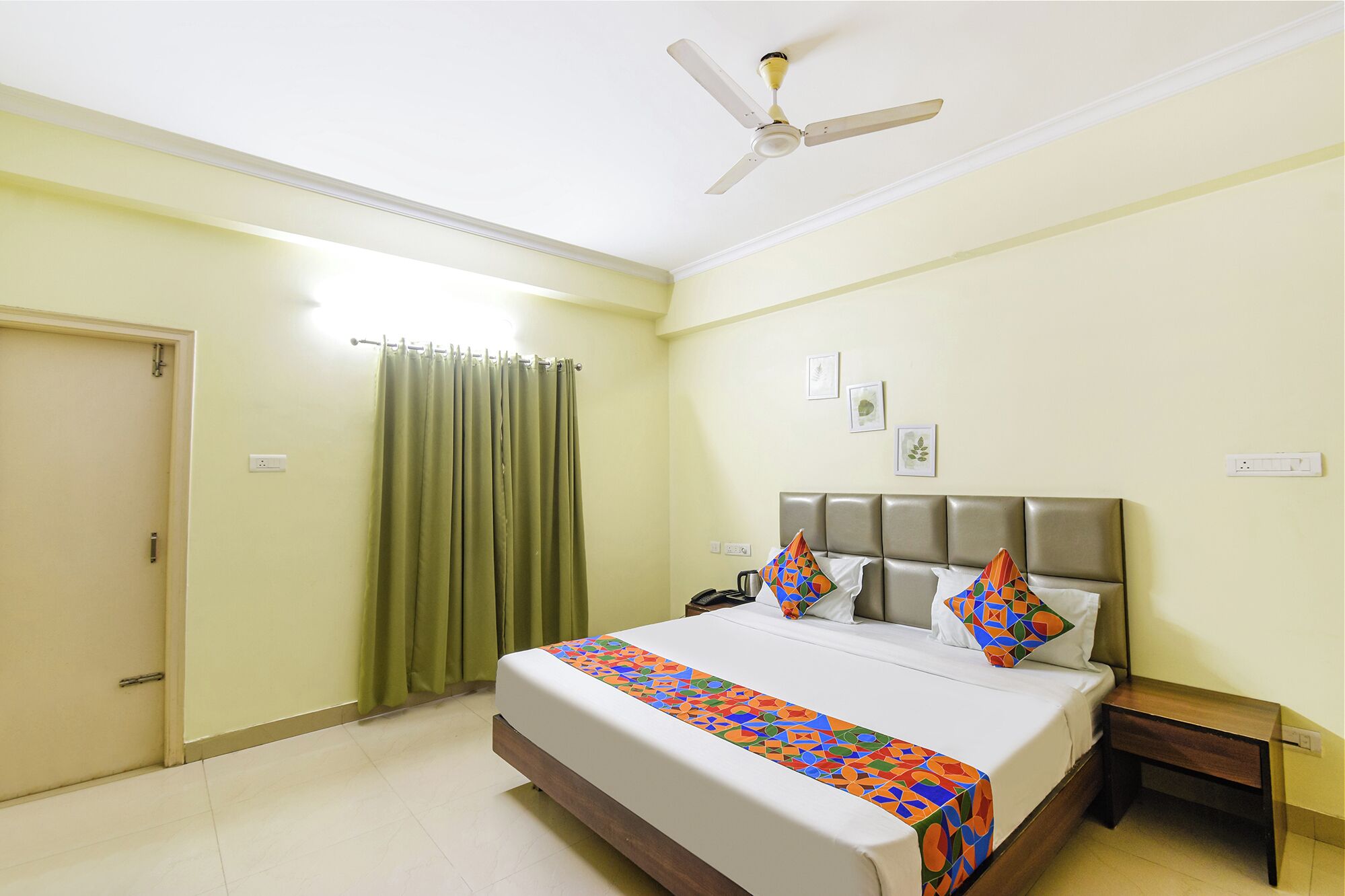 A hotel room with a bed, a nightstand, a ceiling fan, a window with curtains, and a door.