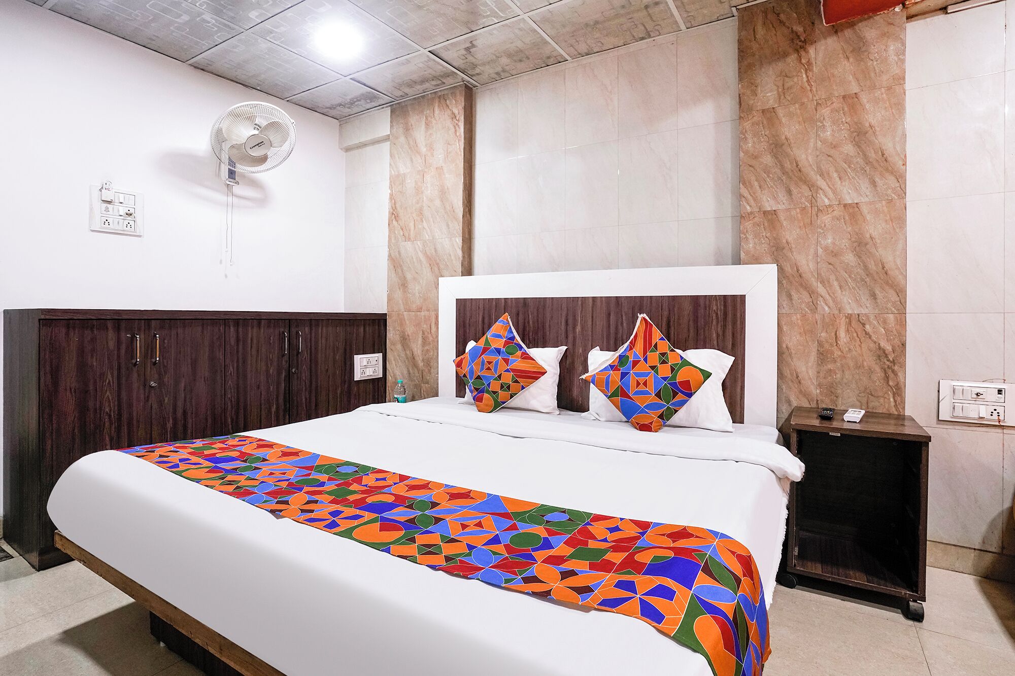 A hotel room with a bed, a nightstand, a wall-mounted fan, and a marble wall.
