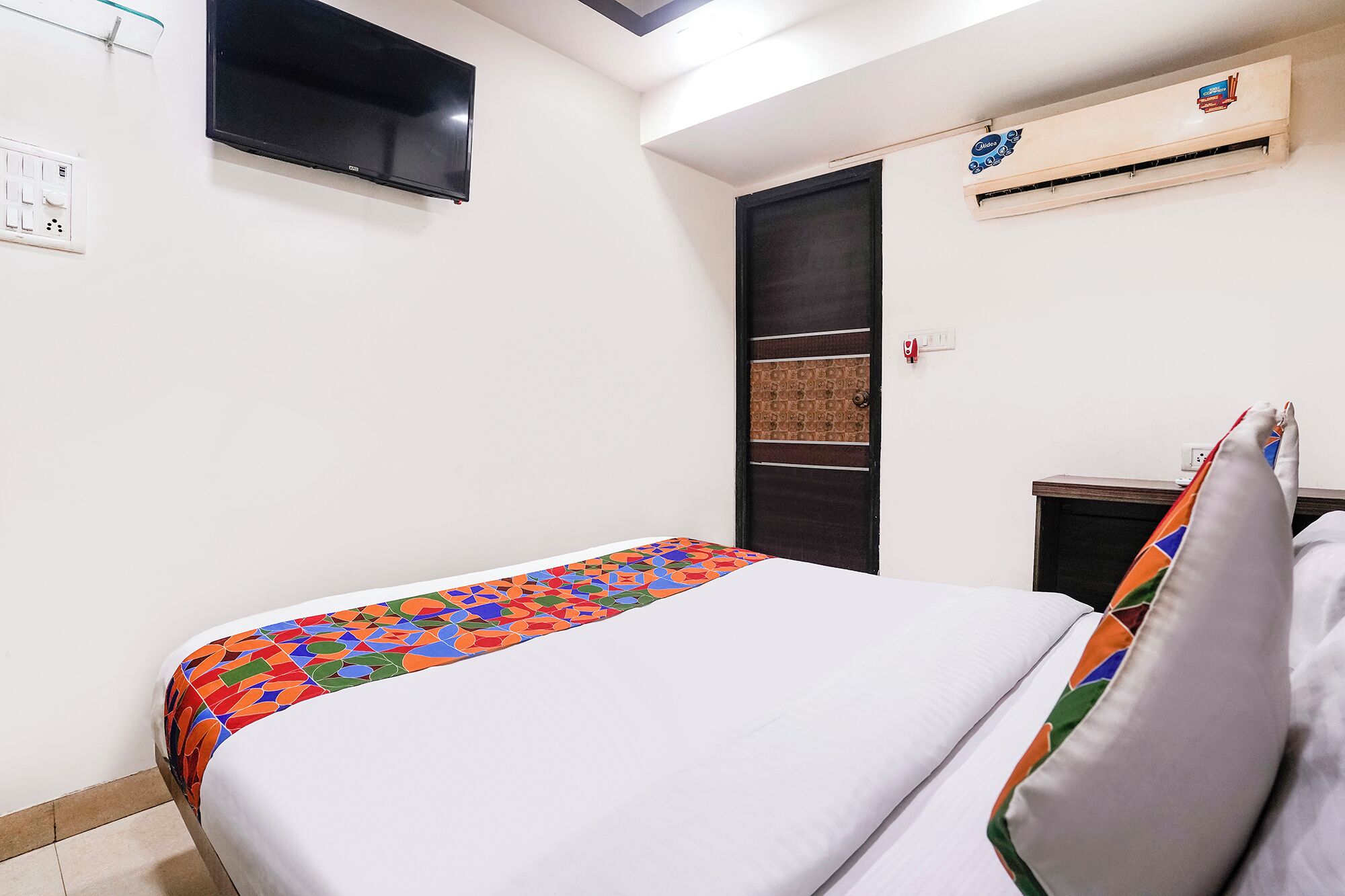 A bedroom with a bed, a wall-mounted air conditioner, a door with a textured panel, and a power outlet.