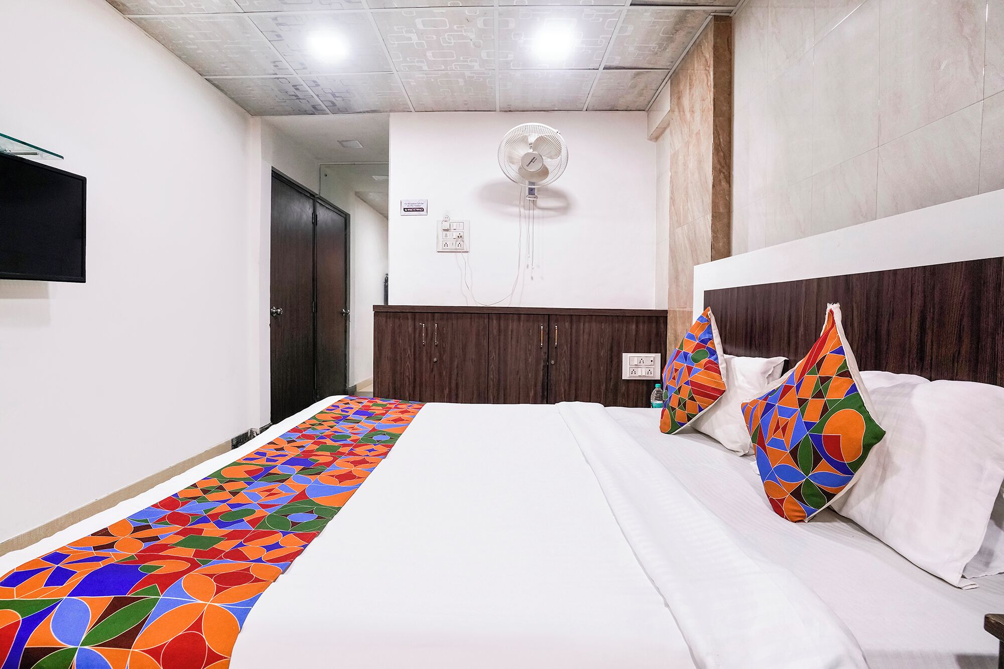 A hotel room with a bed, a wall-mounted fan, a TV, and a door.
