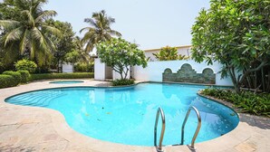 A swimming pool with a curved edge, surrounded by palm trees and a white wall with a stone pattern. - Fabhotel Grand & Resorts (Tirupporur)