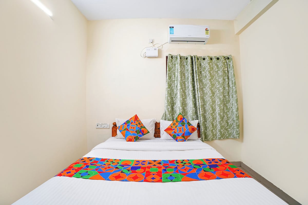 A hotel room with a bed, an air conditioning unit, a power outlet, and a window with curtains.