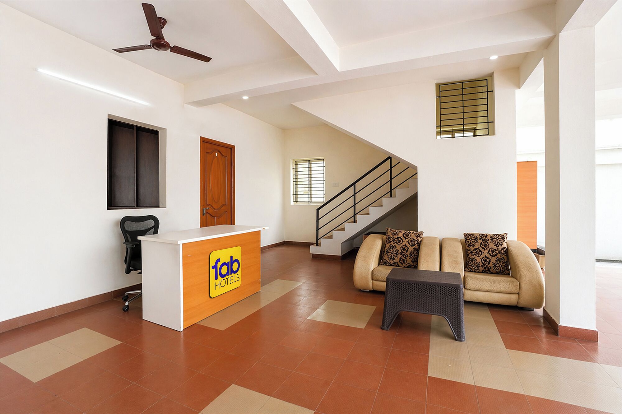 A hotel lobby with a reception desk, a staircase, a sofa, and a ceiling fan.