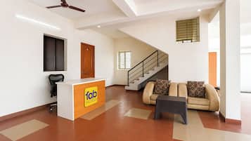 A hotel lobby with a reception desk, a staircase, a sofa, and a ceiling fan.