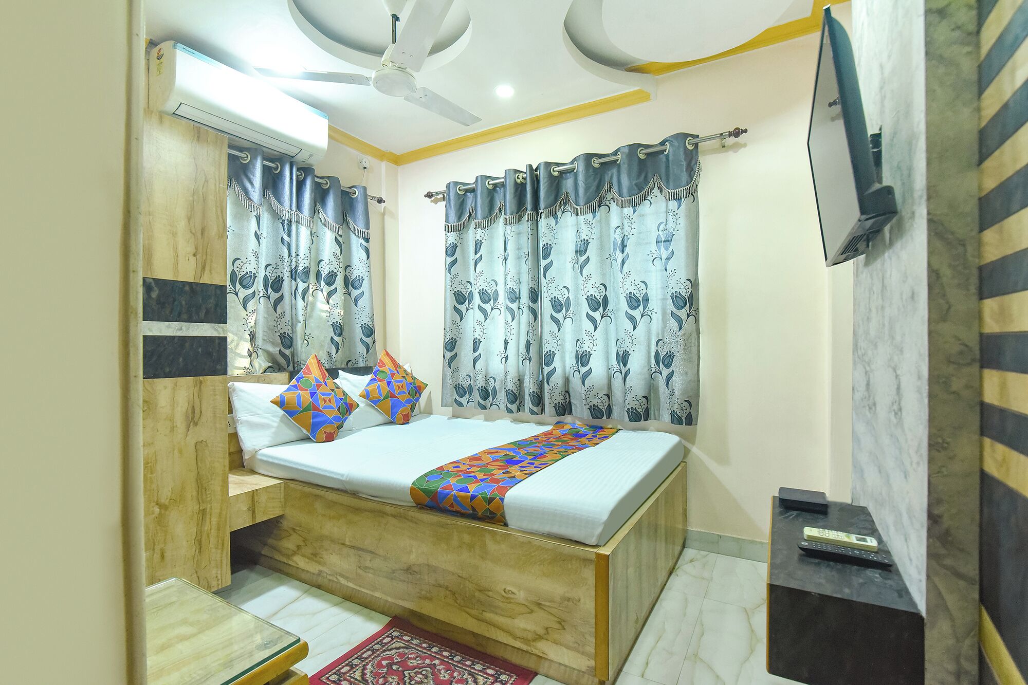 A bedroom with a bed, curtains, a ceiling fan, a TV, and a small table.