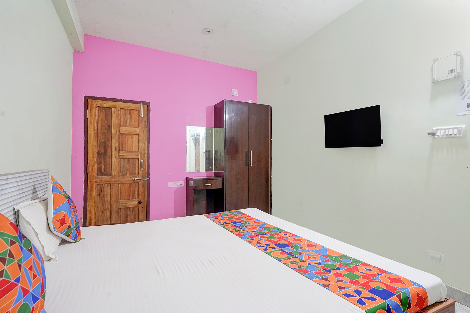A bedroom with a bed, a wooden wardrobe, a pink wall, and a television mounted on the wall.
