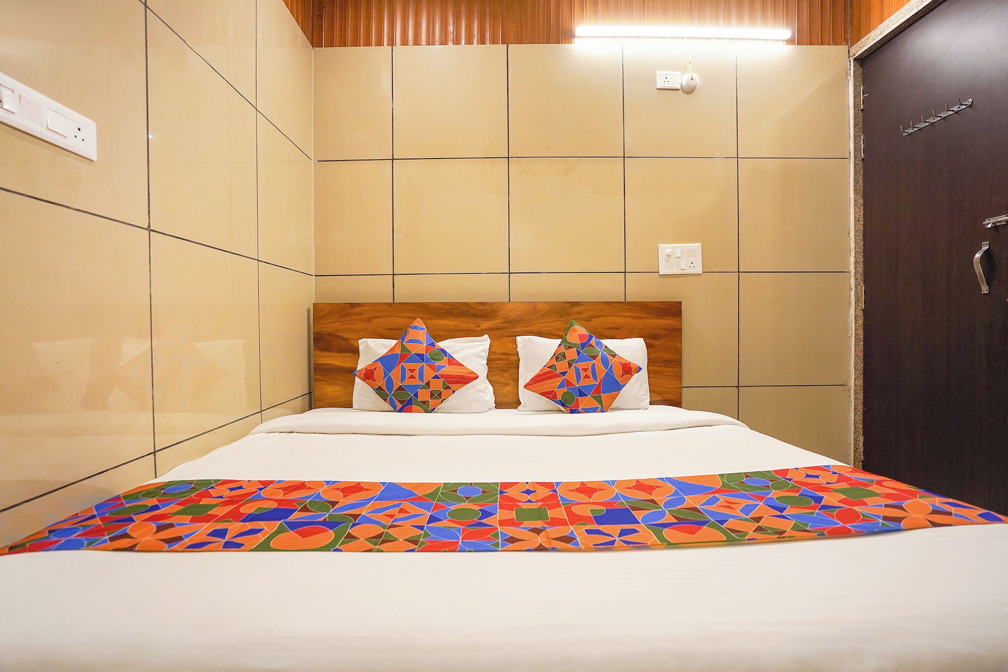 A hotel room with a bed, a wooden headboard, two pillows, a patterned bedspread, and a dark door.