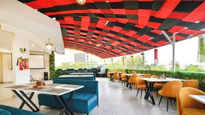 An outdoor dining area with blue and orange seating, a red and black patterned ceiling, and a view of greenery. - Fabhotel The Muffet (Indore)