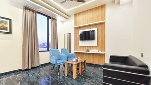 A hotel room with a large window, a flat-screen TV mounted on the wall, a blue armchair, a wooden side table, and a black sofa. - Fabhotel The Nine Prime (Jaipur)
