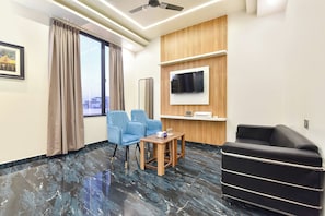 Une chambre d’hôtel dotée d’une grande fenêtre, d’un téléviseur à écran plat fixé au mur, d’un fauteuil bleu, d’une table d’appoint en bois et d’un canapé noir.