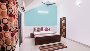 A bedroom with a bed, a ceiling fan, a wall-mounted air conditioner, a patterned curtain, and a tiled floor. - FabHotel Triple Beach House - Nr. Benaulim Beach (Morjim)