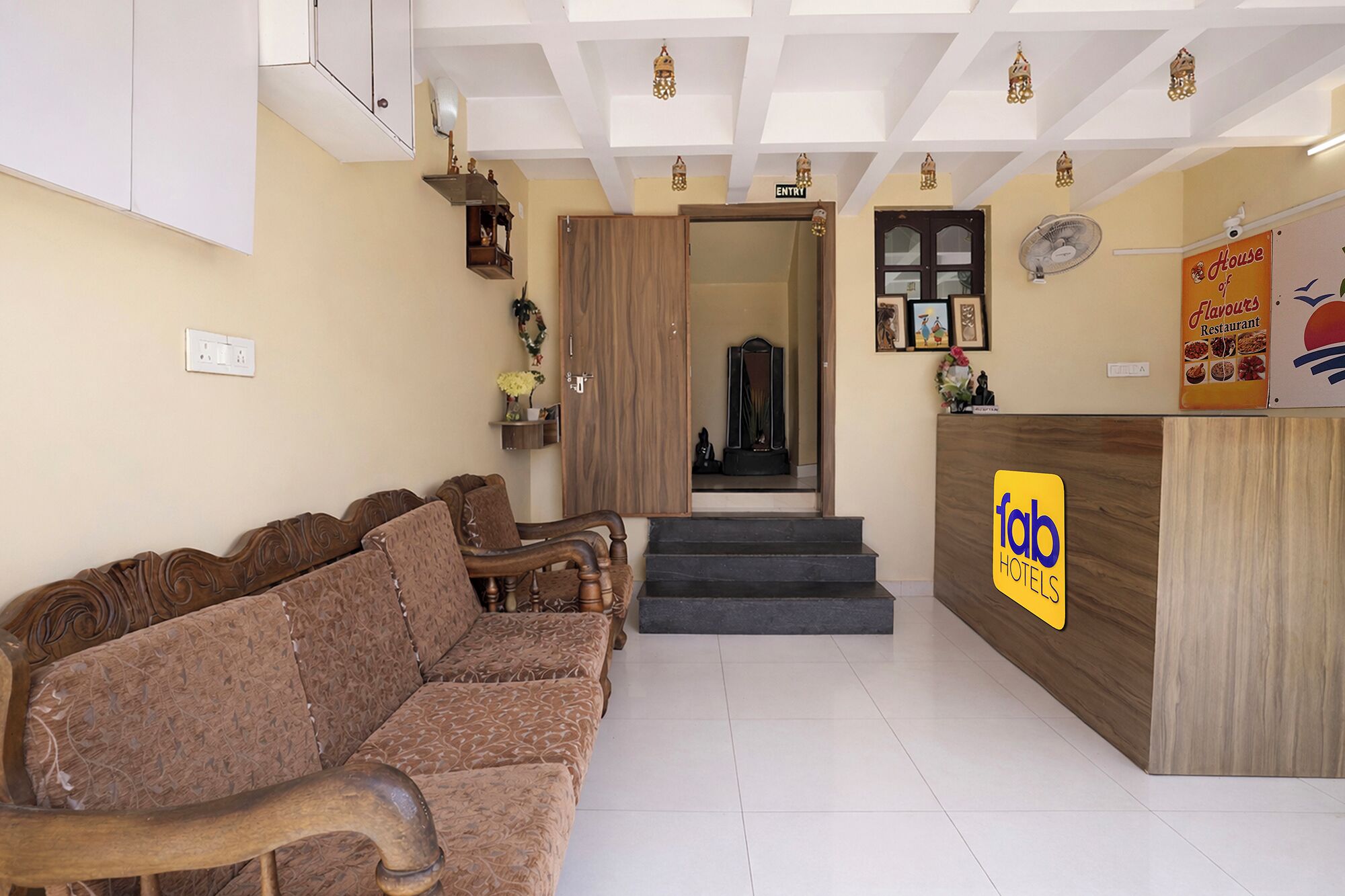 A hotel reception area with a wooden reception desk, a sofa, a mirror, and a staircase leading to an upper floor.