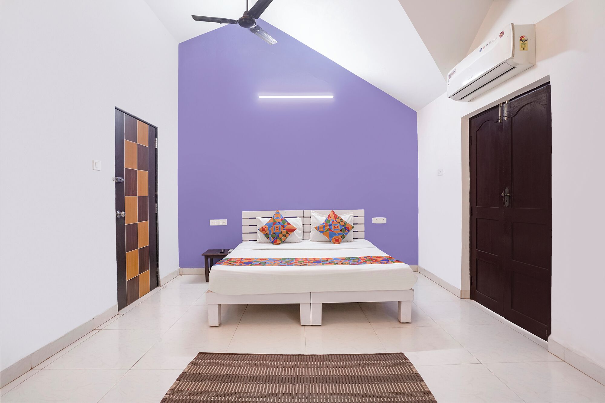A bedroom with a bed, a ceiling fan, an air conditioner, a door, and a wall with a geometric pattern.
