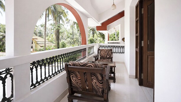 A covered balcony with a view of palm trees and a building, wooden chairs, and a white railing. - FabHotel Triple Beach House - Nr. Benaulim Beach (Morjim)