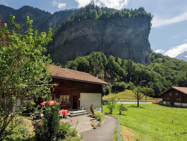 A wooden cabin with a tiled roof, surrounded by greenery and a mountain backdrop.