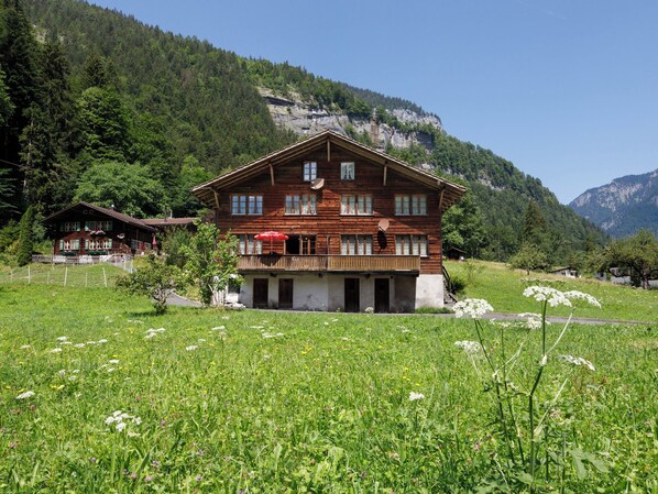 A two-story wooden chalet with a balcony, surrounded by a lush green meadow and mountains in the background.