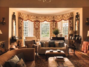 Ein traditionell eingerichteter Raum mit einer Couch, Sesseln, einem Klavier und einem großen Fenster mit Vorhängen.