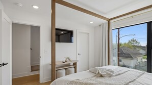 Ein Schlafzimmer mit einem Bett, einem Nachttisch, einem Spiegel und einem Fenster mit Blick auf Häuser.