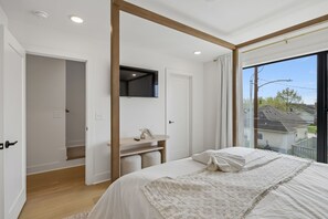 Ein Schlafzimmer mit einem Bett, einem Nachttisch, einem Spiegel und einem Fenster mit Blick auf Häuser.