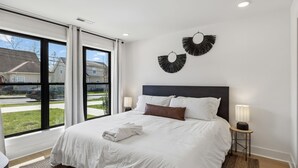 A bedroom with a large bed, two wall-mounted lamps, and a view of a street through a large window. - Upscale home featuring rooftop lounge with hottub! (Nashville)