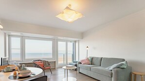 Un salon moderne comprenant un canapé, une table à manger avec des chaises, une table basse et une vue sur la mer grâce à de grandes fenêtres.