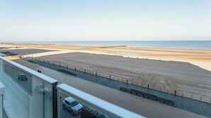Une vue panoramique sur la côte, prise depuis un point d’observation élevé, qui offre un paysage comprenant une plage de sable, une promenade dallée et une étendue d’eau visible au loin.