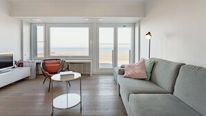 Un salon moderne avec un canapé, une petite table et un fauteuil, de grandes fenêtres donnant sur la plage et un lampadaire.