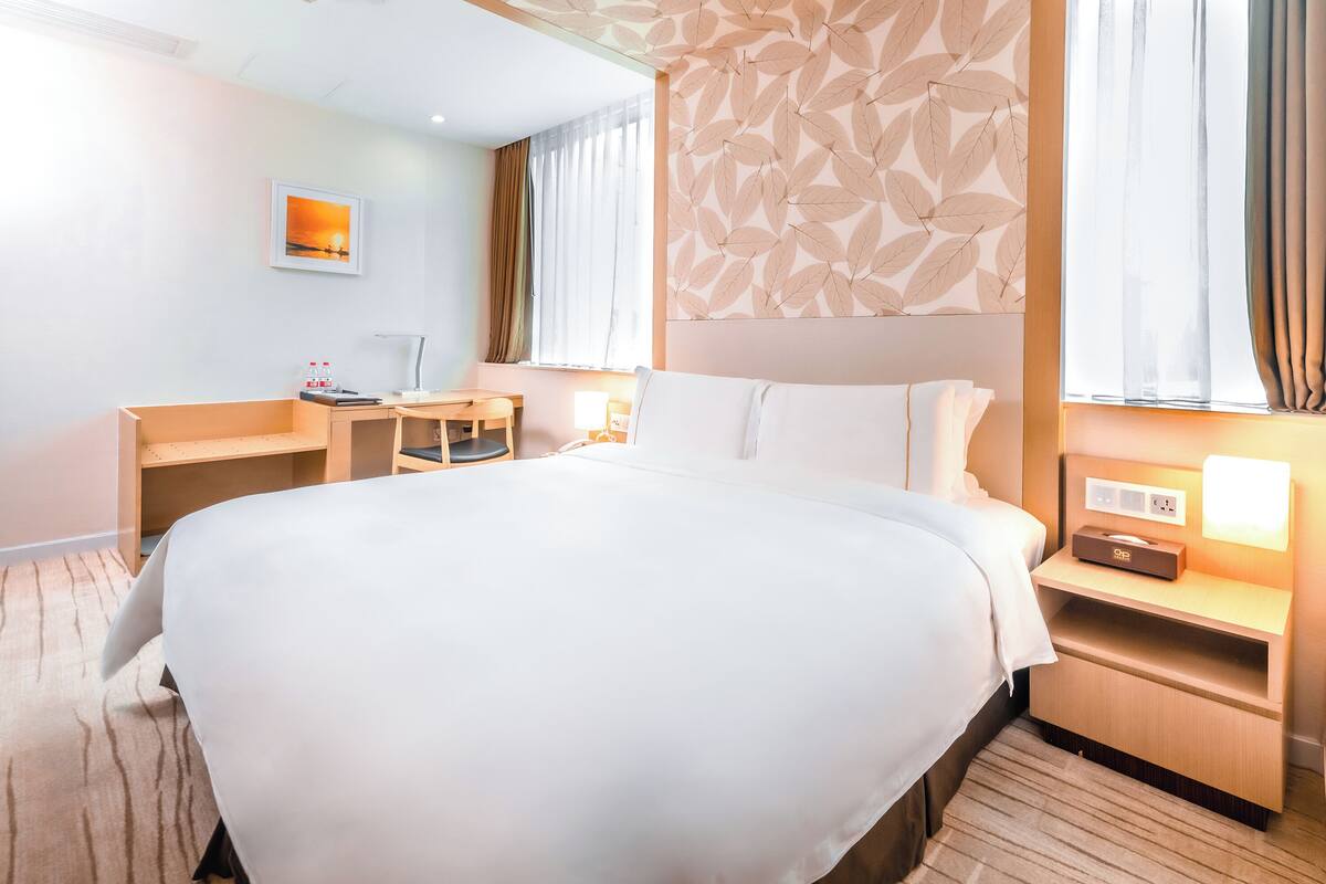 Habitación de hotel con una cama grande, un escritorio, una silla, una mesita de noche con lámpara y un enchufe en la pared.