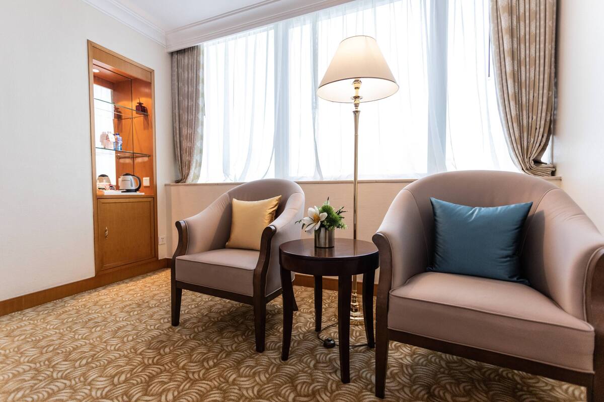 Habitación de hotel con dos sillones, una mesita de noche con lámpara, un armario y una alfombra estampada.